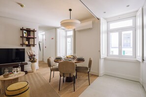 A modern living room with a television, dining area with a round table and chairs, a sofa, and a large window providing natural light. - Ando Living - Tejo Palace (Lisboa)
