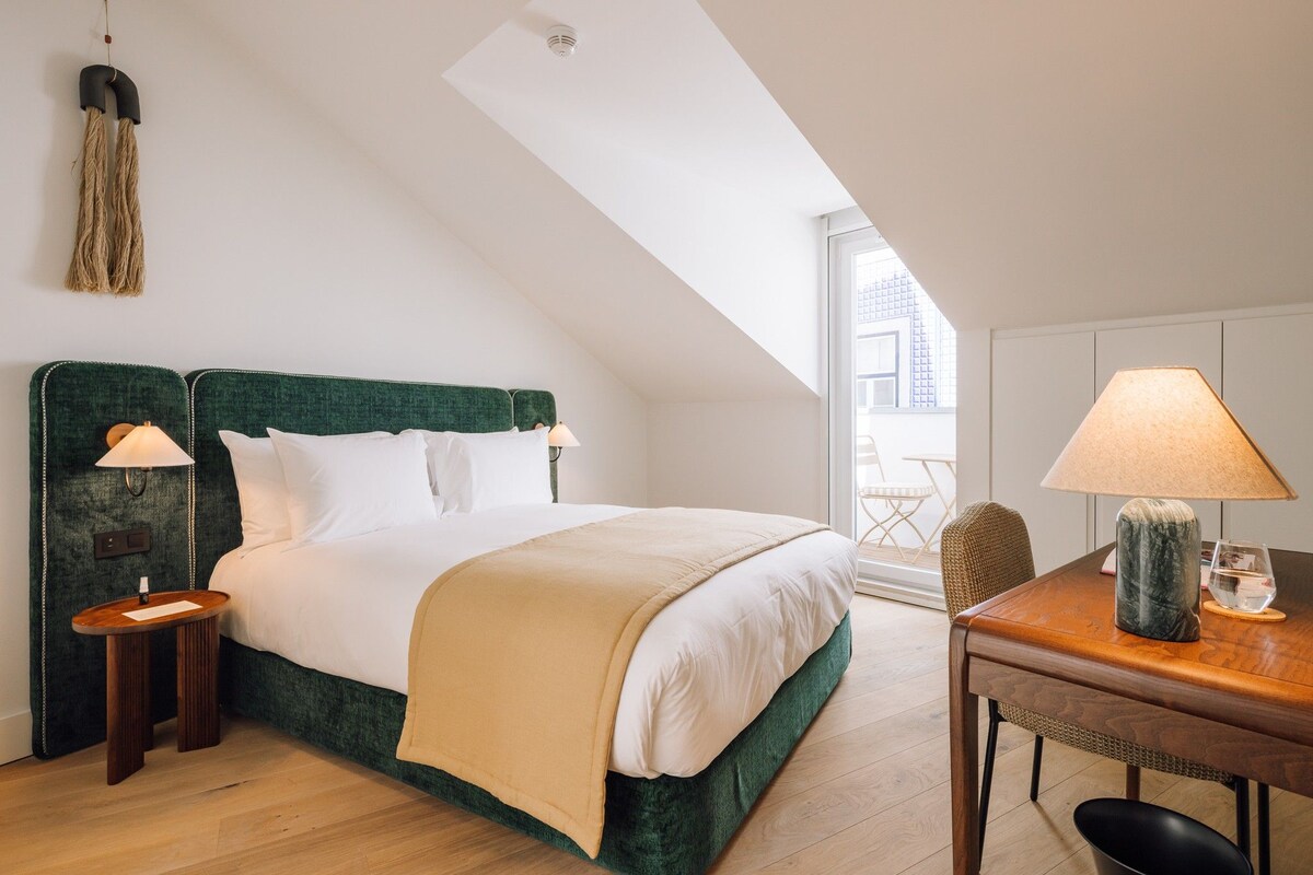 A bedroom with a bed, bedside tables, a lamp, a chair, a table, and a view of the outside.