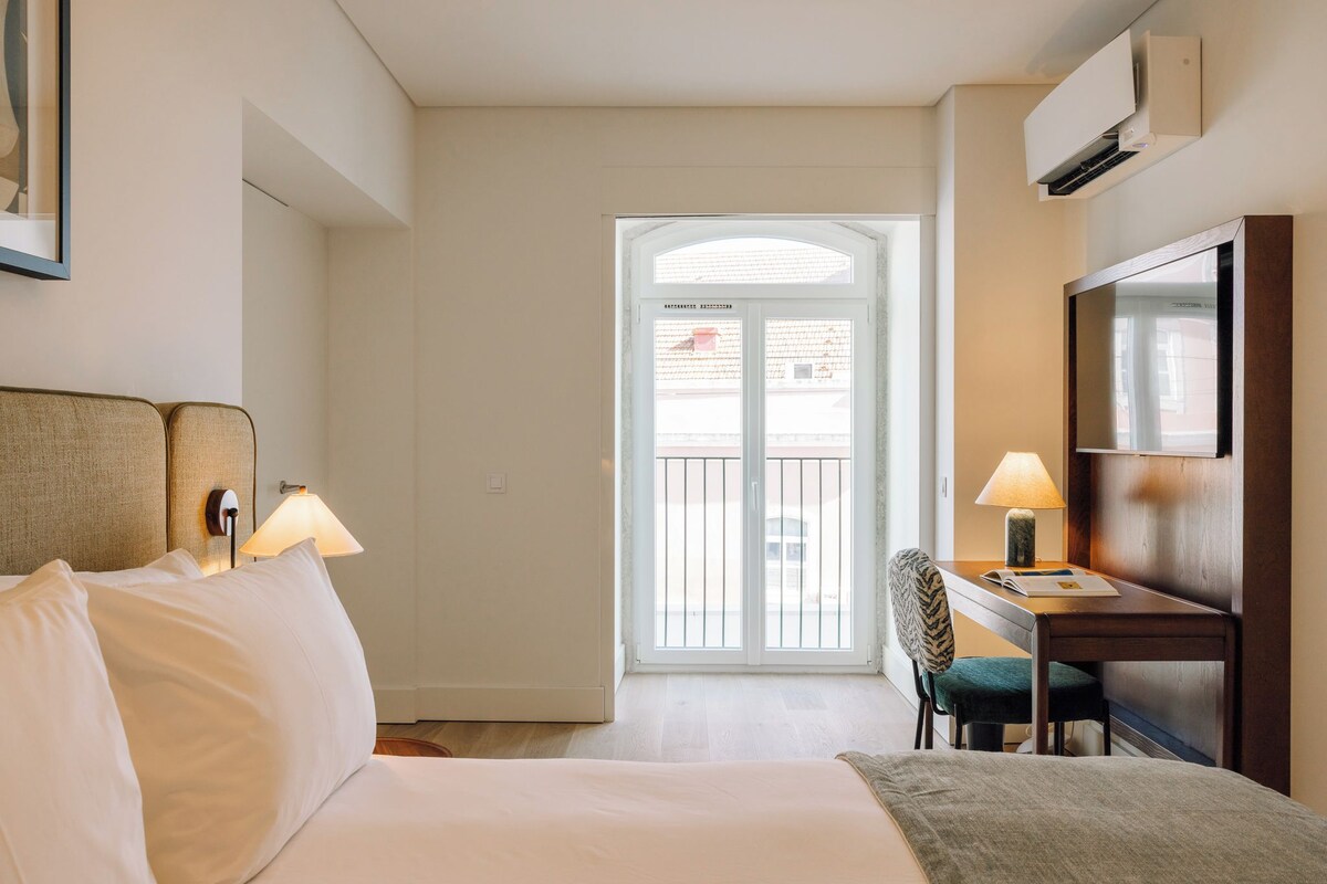 A hotel room with a bed, two bedside lamps, a desk with a chair, a TV, and a large window with a view of the outside.