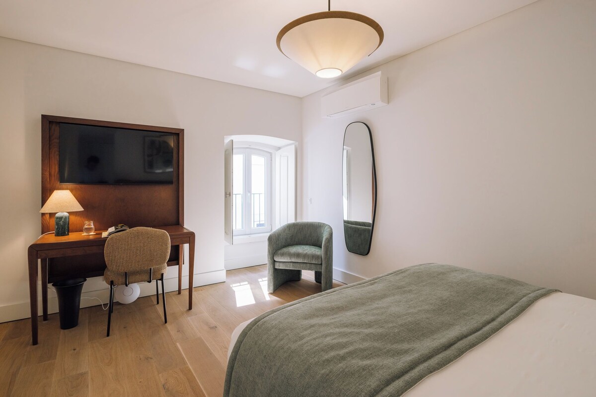 A hotel room with a bed, a desk with a lamp, a chair, a large mirror, and a window.