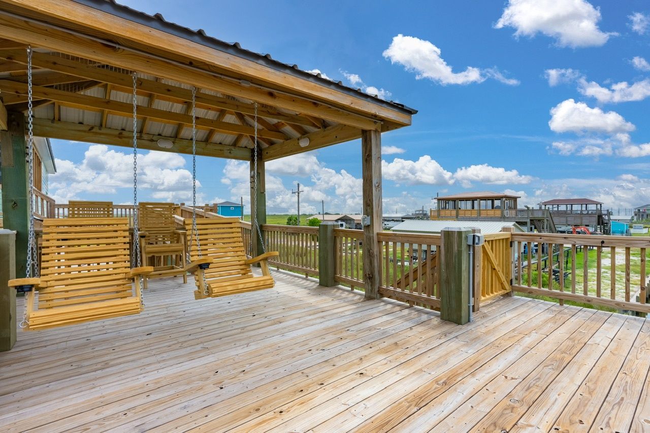 Una terraza de madera con un conjunto de columpios y un área techada, con vista a un cuerpo de agua y edificios.