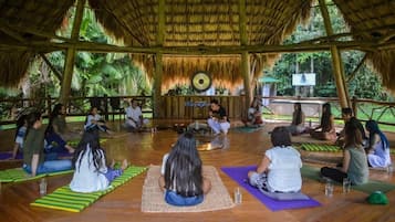 A group of people sitting on mats in a circular arrangement, facing a central speaker, in an open-air setting with a thatched roof and wooden beams.