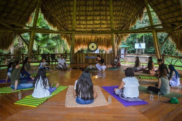 A group of people sitting on mats in a circular arrangement, facing a central speaker, in an open-air setting with a thatched roof and wooden beams.