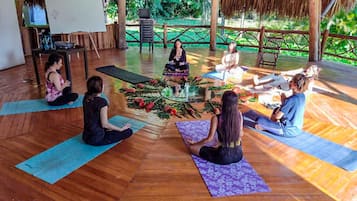 Eine Gruppe von Menschen sitzt auf Yogamatten in einem Raum mit Holzboden und einem Strohdach.