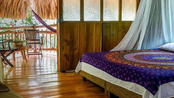 A room with a bed, wooden flooring, a thatched roof, and a view of greenery.