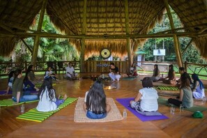 A group of people sitting on mats in a circular arrangement, facing a central speaker, in an open-air setting with a thatched roof and wooden beams.