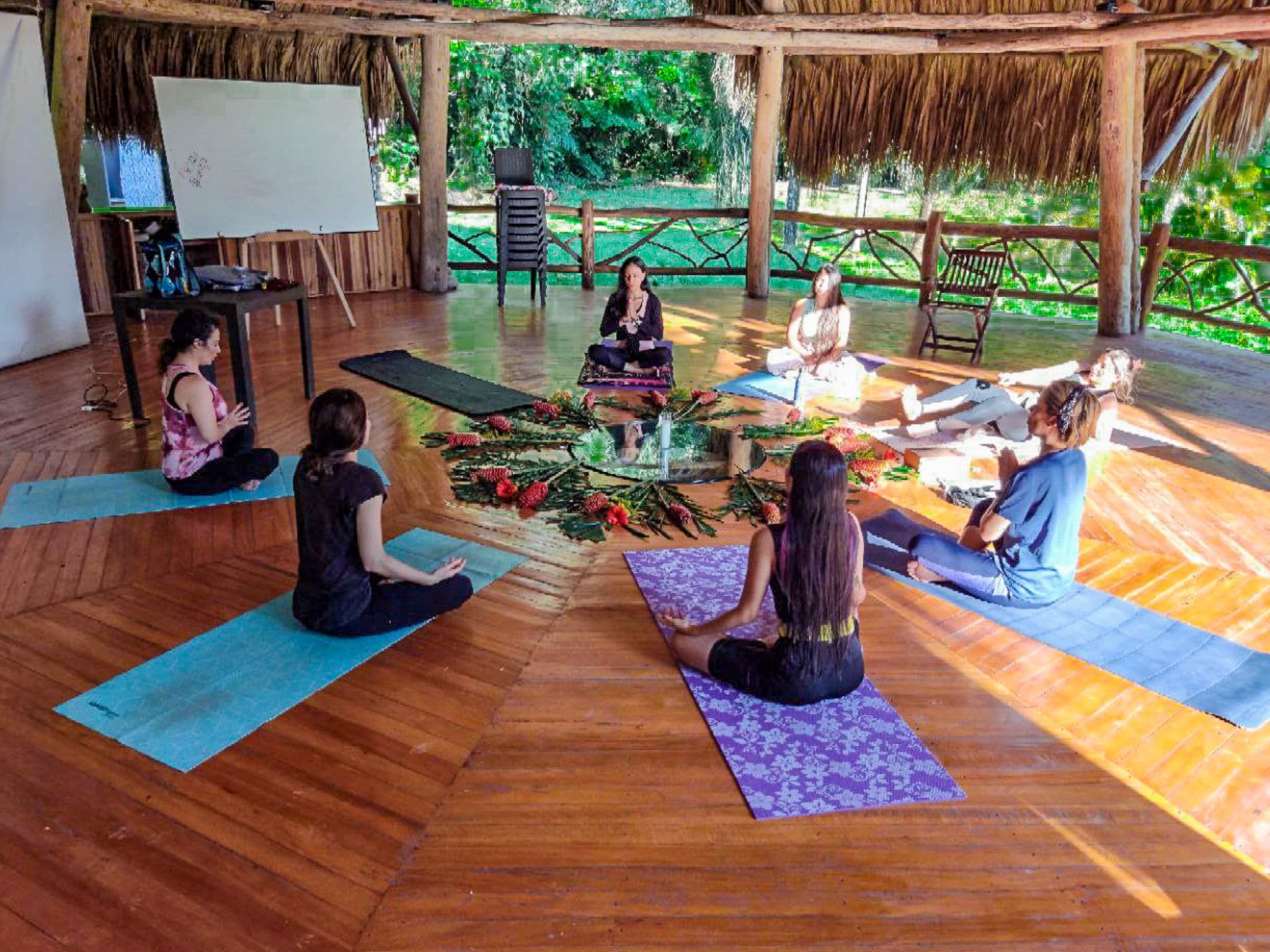 Eine Gruppe von Menschen sitzt auf Yogamatten in einem Raum mit Holzboden und einem Strohdach.