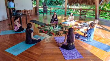 Eine Gruppe von Menschen sitzt auf Yogamatten in einem Raum mit Holzboden und einem Strohdach.