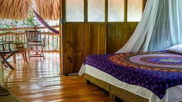 Una habitación con una cama, piso de madera, techo de paja y vista a zonas verdes.