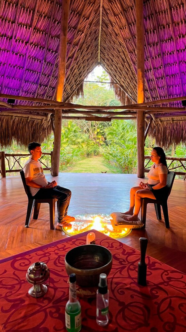 Two people seated at a table with a fire in the center, under a thatched roof with a view of greenery.