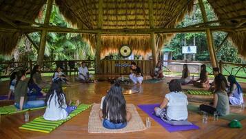 A group of people sitting on mats in a circular arrangement, facing a central speaker, in an open-air setting with a thatched roof and wooden beams.