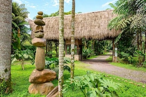 Una estructura con techo de paja rodeada de abundante vegetación y una escultura de piedras equilibradas.