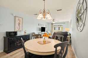 A dining area with a round table, chairs, a vase with flowers, a ceiling fan, and a mounted television. - Grand Strand Little Retreat (Garden City Beach)