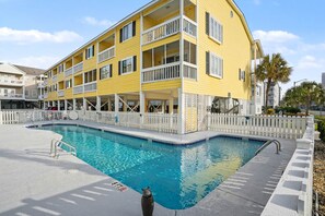 A yellow multi-story building with a swimming pool, white railings, and a covered area with outdoor seating. - Grand Strand Little Retreat (Garden City Beach)