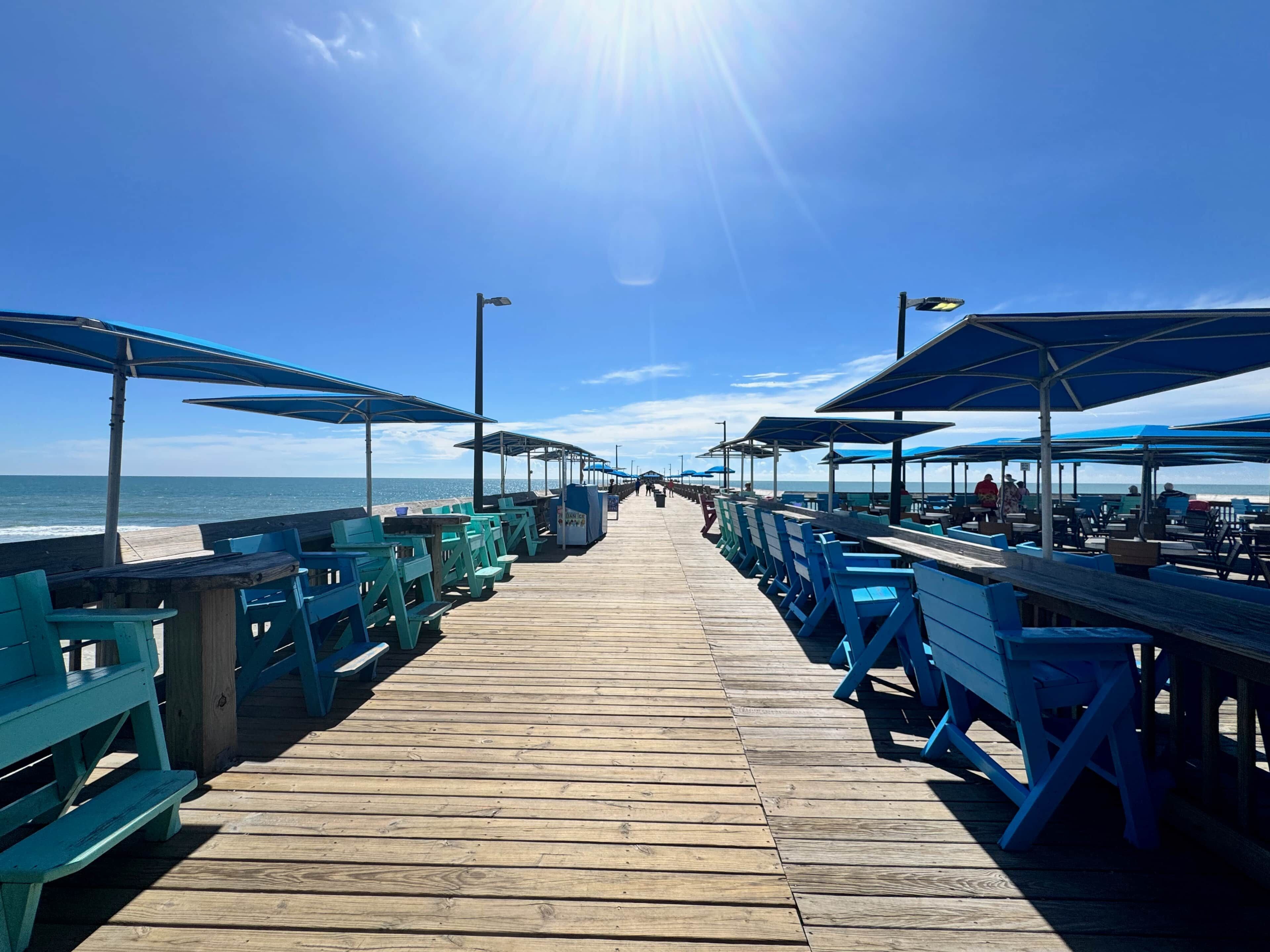 A wooden pier with blue benches and umbrellas, leading to the beach.