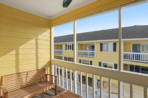A balcony with a wooden bench, a view of a yellow building with multiple windows and balconies, and a clear sky. - Grand Strand Little Retreat (Garden City Beach)