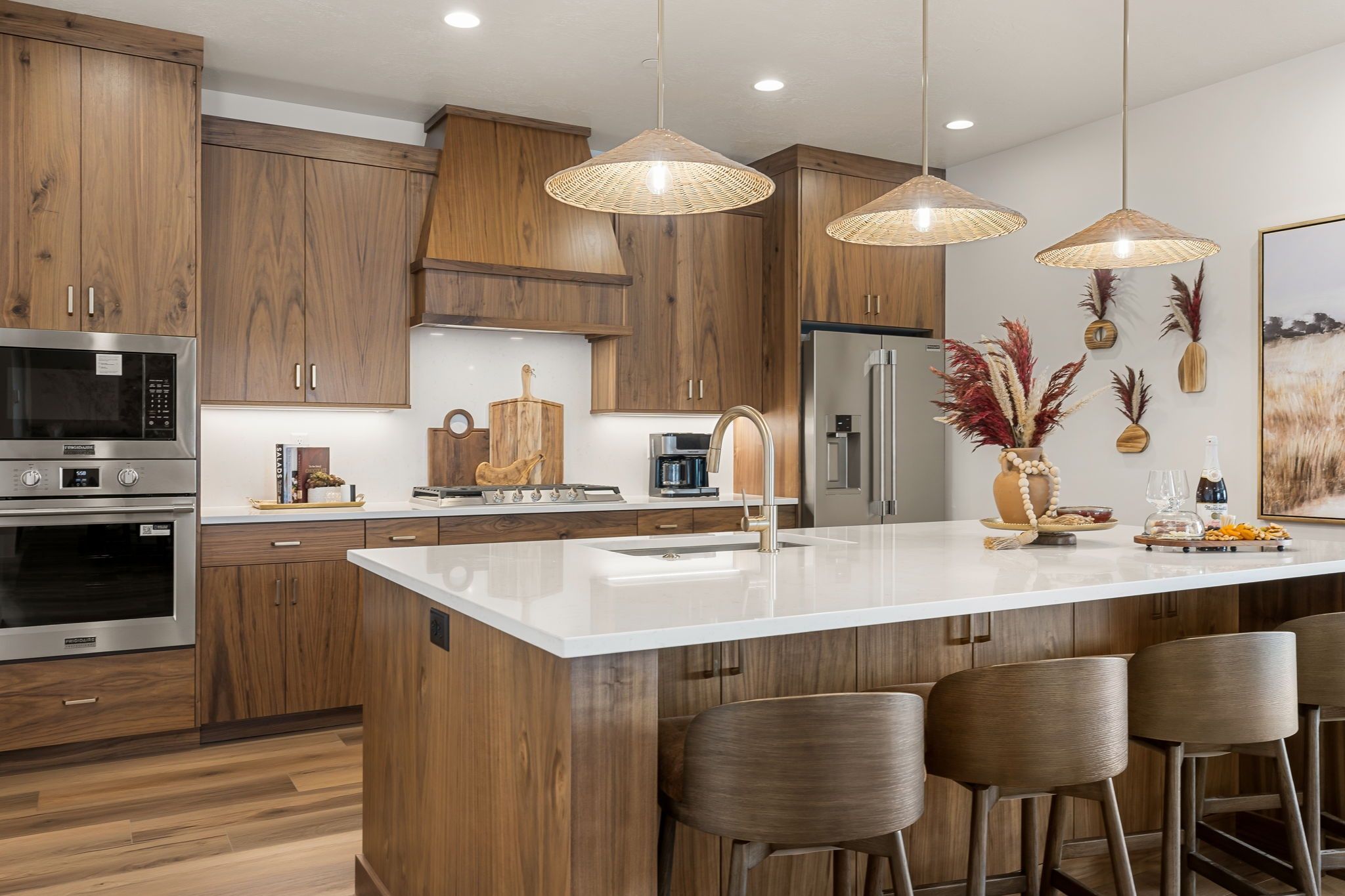 A modern kitchen with wooden cabinets, stainless steel appliances, a central island with bar stools, pendant lighting, and decorative wall art.