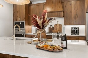 A kitchen island with a food spread, a bottle of wine, and a decorative vase with dried plants. - The Isle Retreat 6 Bedroom Townhouse (Washington)