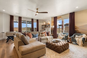 Ein geräumiges Wohnzimmer mit einer Couch, Hocker und Sesseln, einem Deckenventilator und großen Fenstern mit Blick ins Freie.