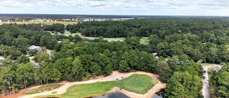 An aerial view of a golf course with a pond, surrounded by dense trees and a clear sky.