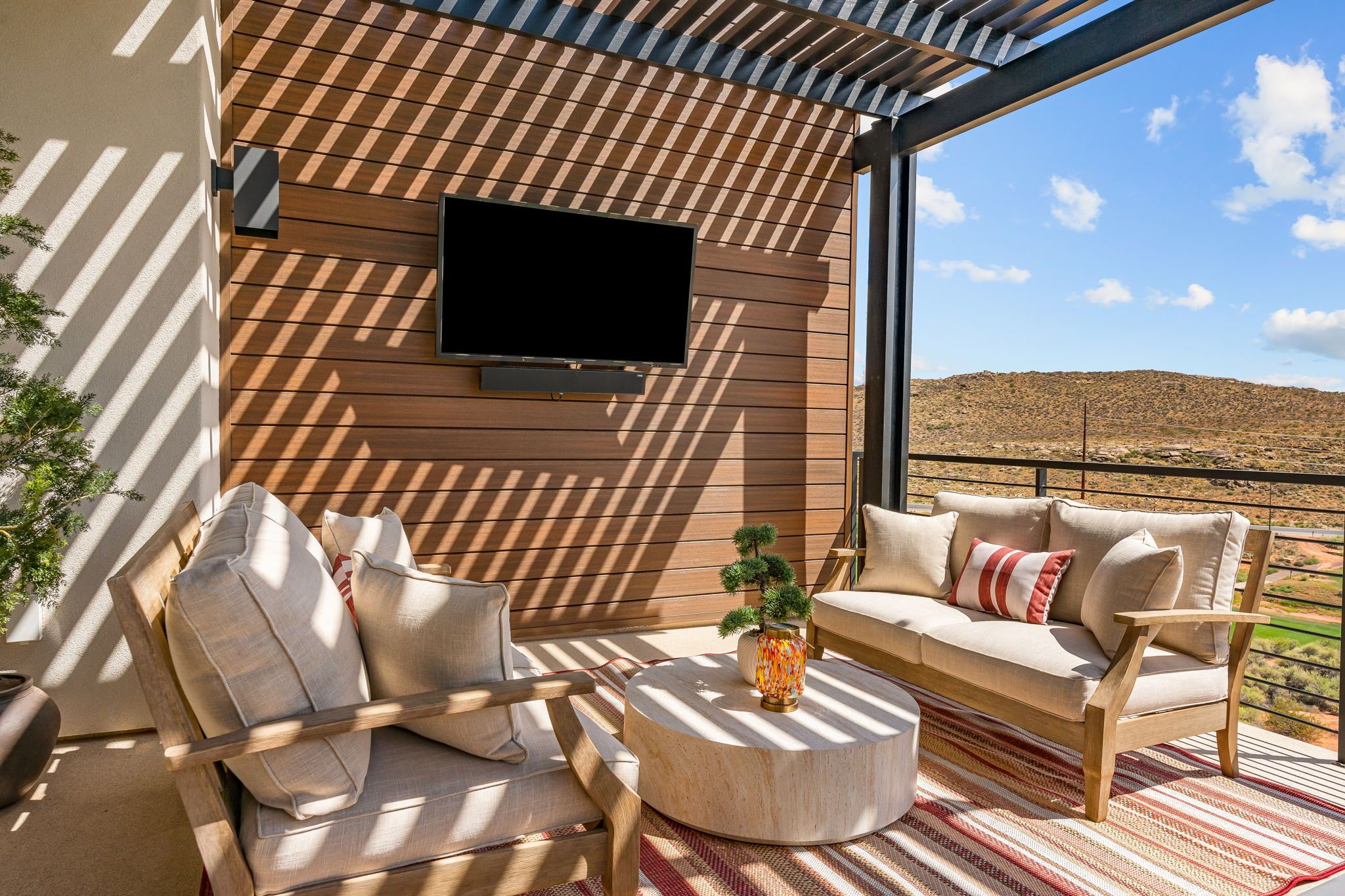 Un espace de détente extérieur moderne, doté d’un téléviseur à écran plat fixé au mur, d’une table ronde en bois et d’un canapé avec des coussins.