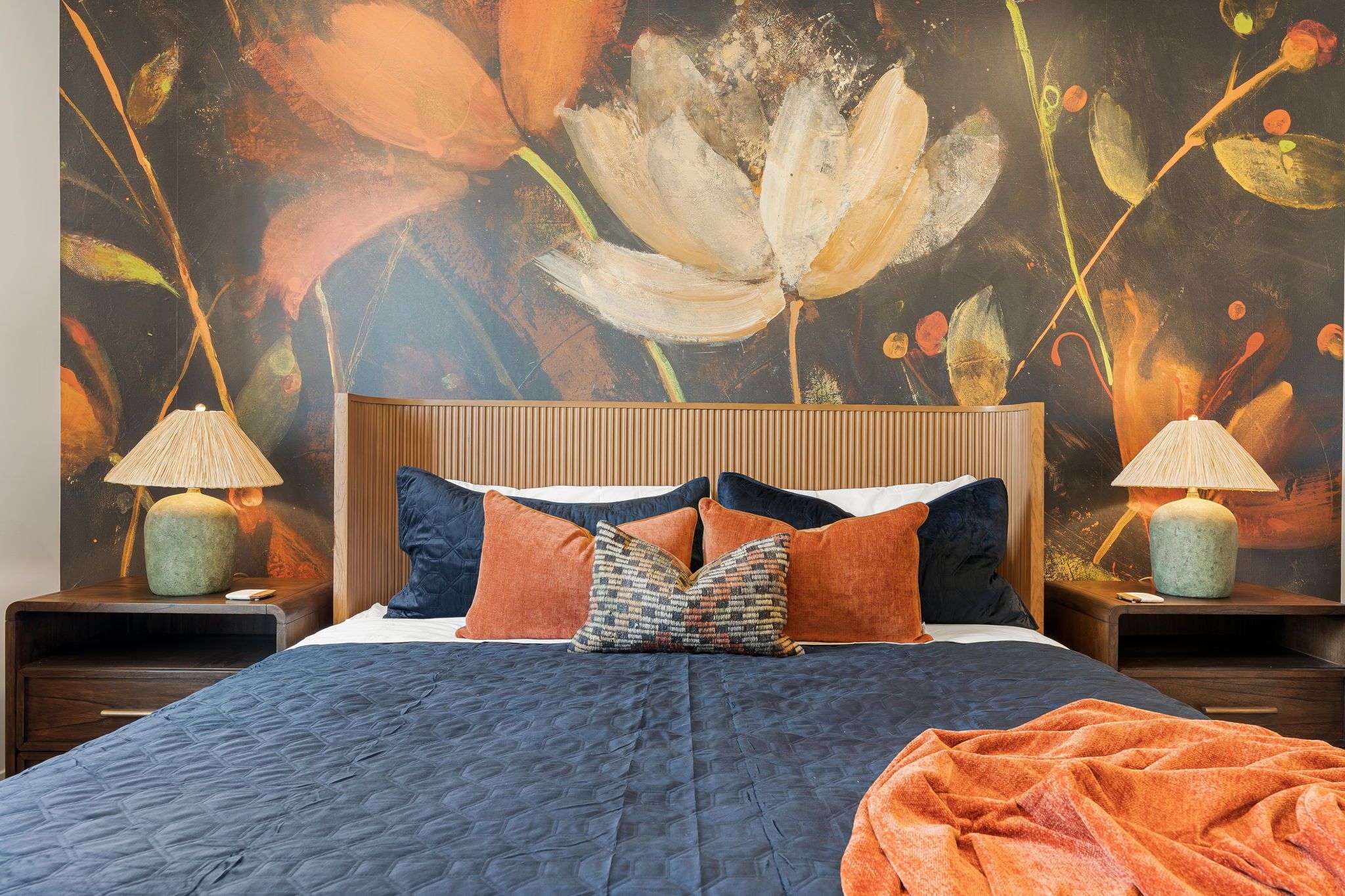 Un lit recouvert d’une couverture sombre, agrémenté de plusieurs oreillers, et un grand motif floral ornant le mur situé derrière.