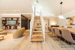 Ein modernes Interieur mit einer Holztreppe, einem Essbereich und einem Wohnraum mit Fernseher und Bücherregalen.