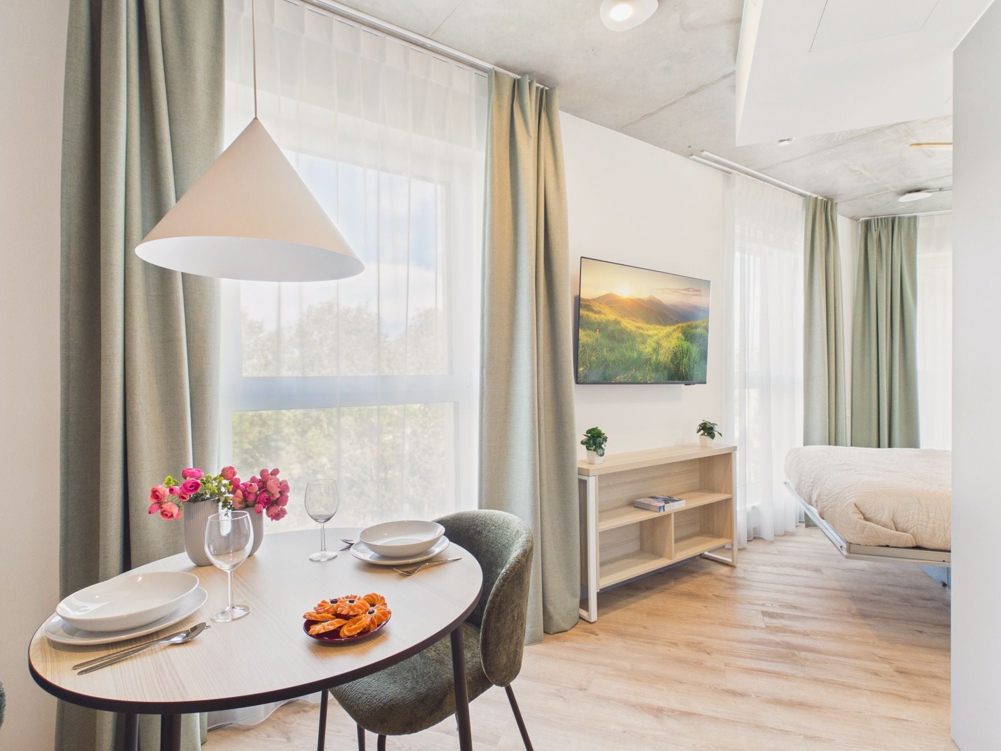 Ein modernes Hotelzimmer mit Essbereich, einem Bett, einem Fernseher und einem großen Fenster mit Vorhängen.