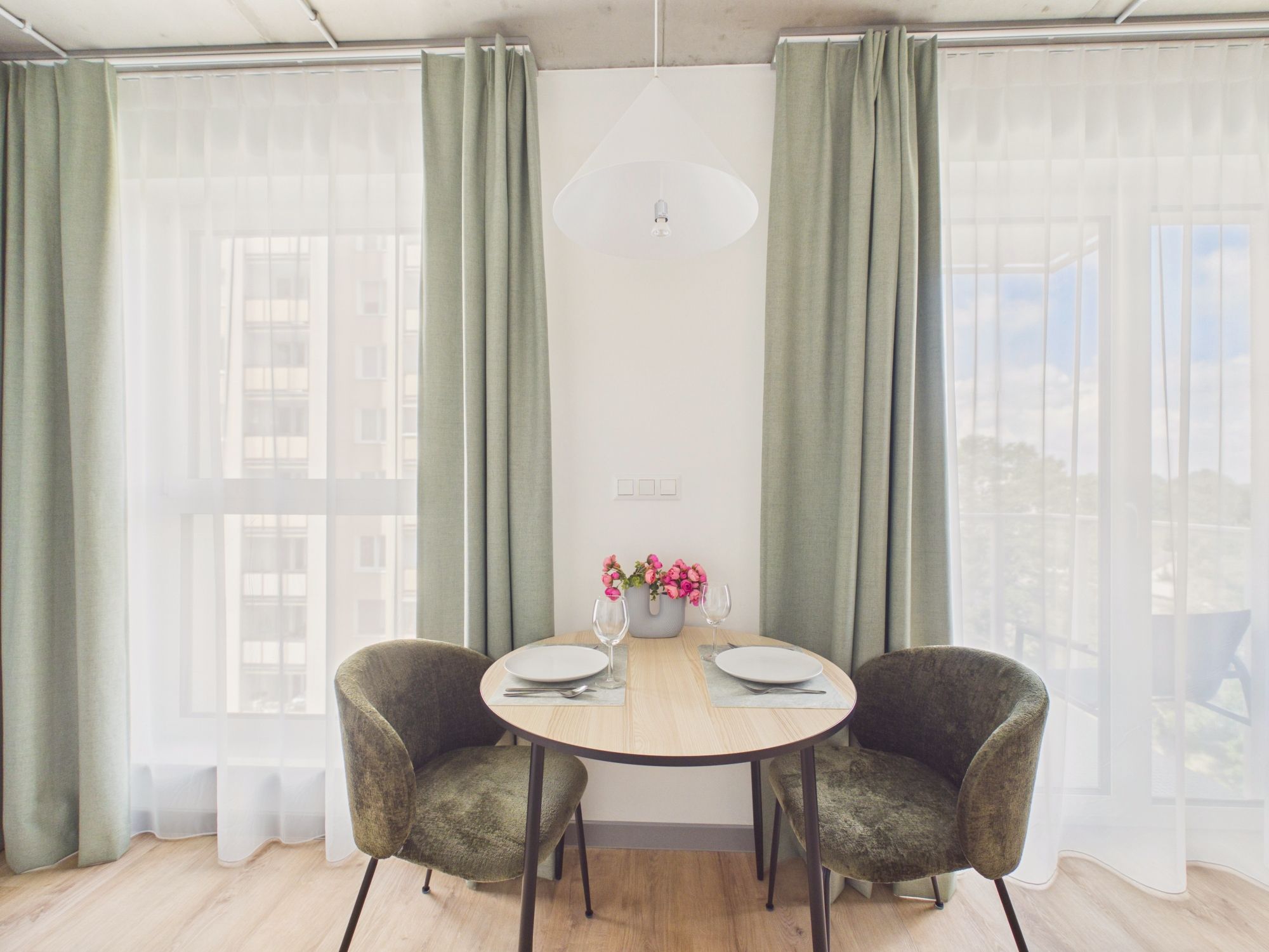 A dining area with a round table, two chairs, a vase of flowers, and a view of buildings through sheer curtains.