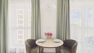 A dining area with a round table, two chairs, a vase of flowers, and a view of buildings through sheer curtains.