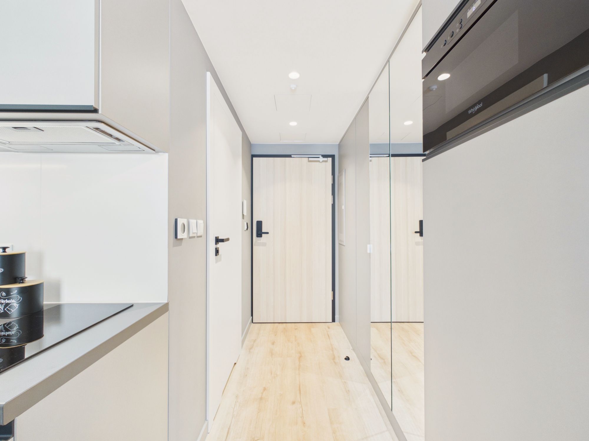 A narrow hallway with a wooden floor, a door at the end, and a kitchen area on the left with a black countertop and a stove.