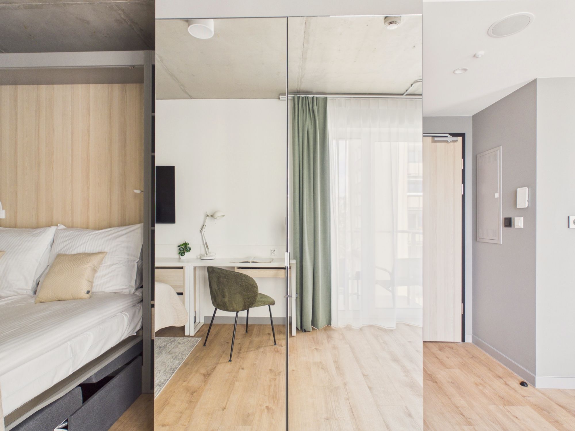 Ein modernes Schlafzimmer mit Bett, Schreibtisch, Stuhl und Blick auf eine Stadtlandschaft durch ein Fenster.