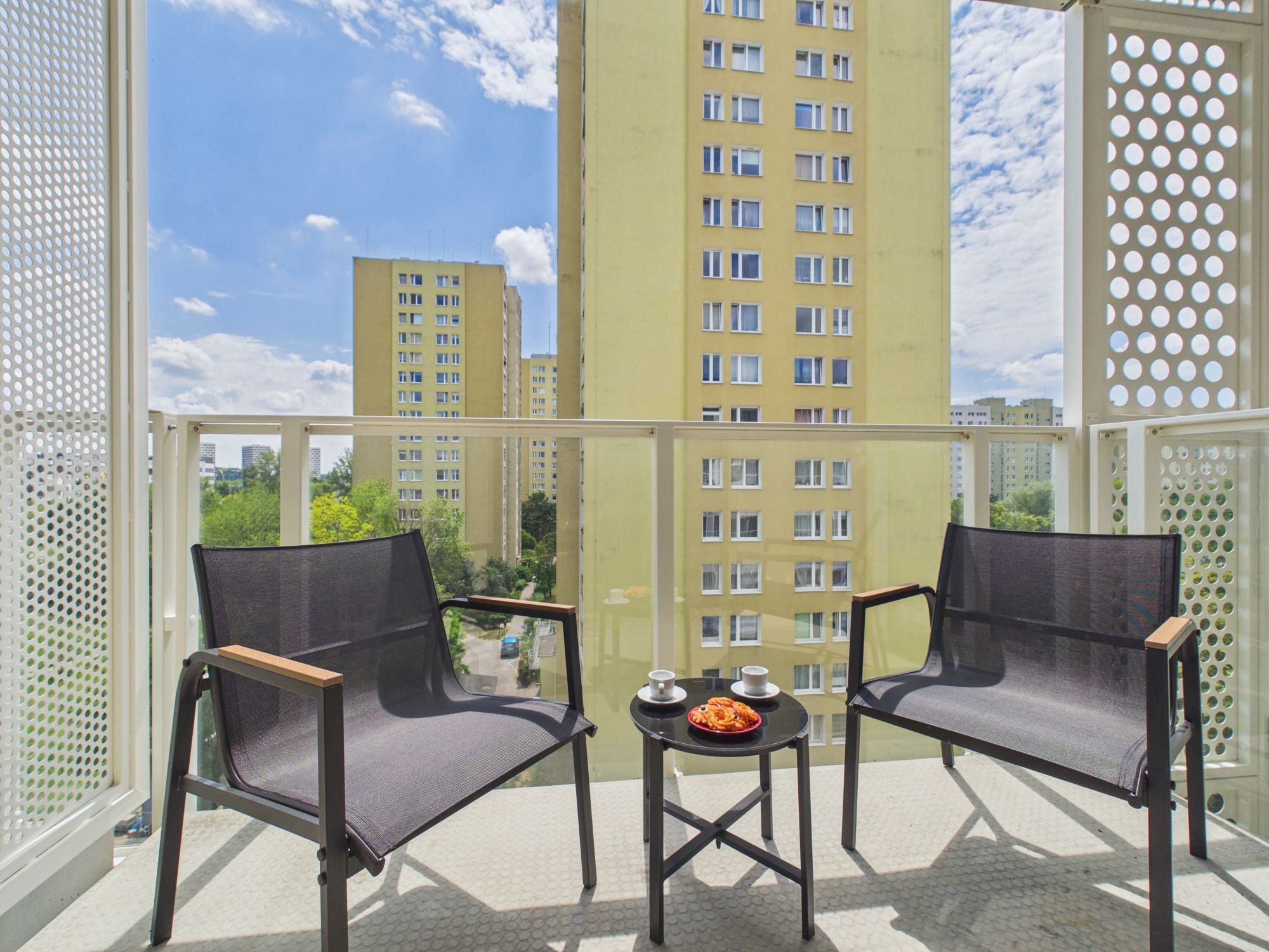 A balcony with two chairs and a small table, overlooking a cityscape with high-rise buildings.