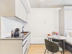 A compact kitchenette with a stove, sink, and modern appliances. There is a dining area with a table and chairs, and a bed with a white headboard. - NanoApart by Solarento (Warszawa)