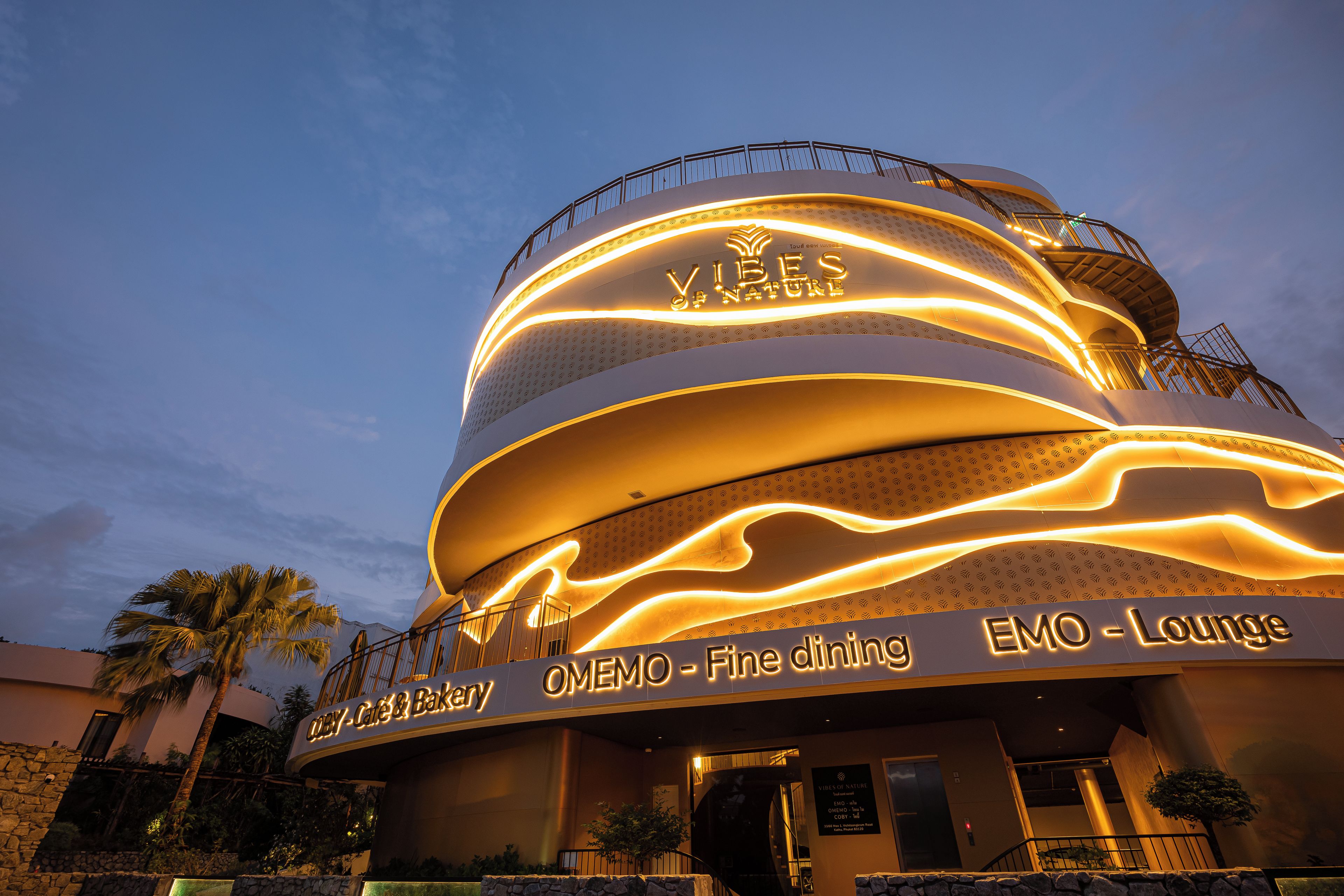 Un edificio con fachada curva, iluminación de letreros que indican 'Vibes of Nature' y 'EMO Lounge', y un cartel que dice 'Odey - Café & Panadería'.