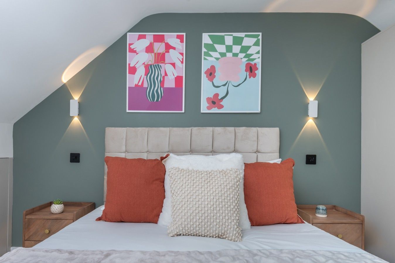 A bedroom with a bed, two pillows, a headboard, two wall-mounted lights, and two framed artworks.