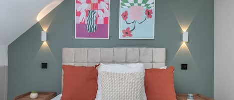 Un dormitorio con cama, dos almohadas, cabecera, dos lámparas fijadas a la pared y dos obras de arte enmarcadas.