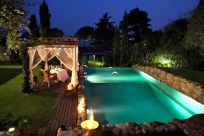 Un área de piscina con terraza de madera, zona de estar cubierta y pared de piedra.