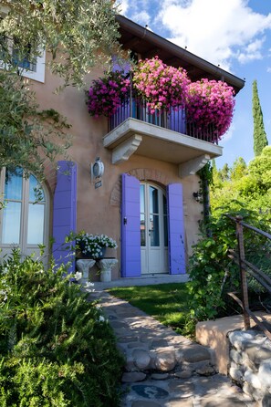 Una casa con balcón adornado con flores rosadas, un camino de piedra que conduce a la entrada y un jardín bien cuidado.