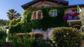 Ein zweistöckiges Gebäude mit Balkon, umgeben von üppigem Grün und blühenden Pflanzen.