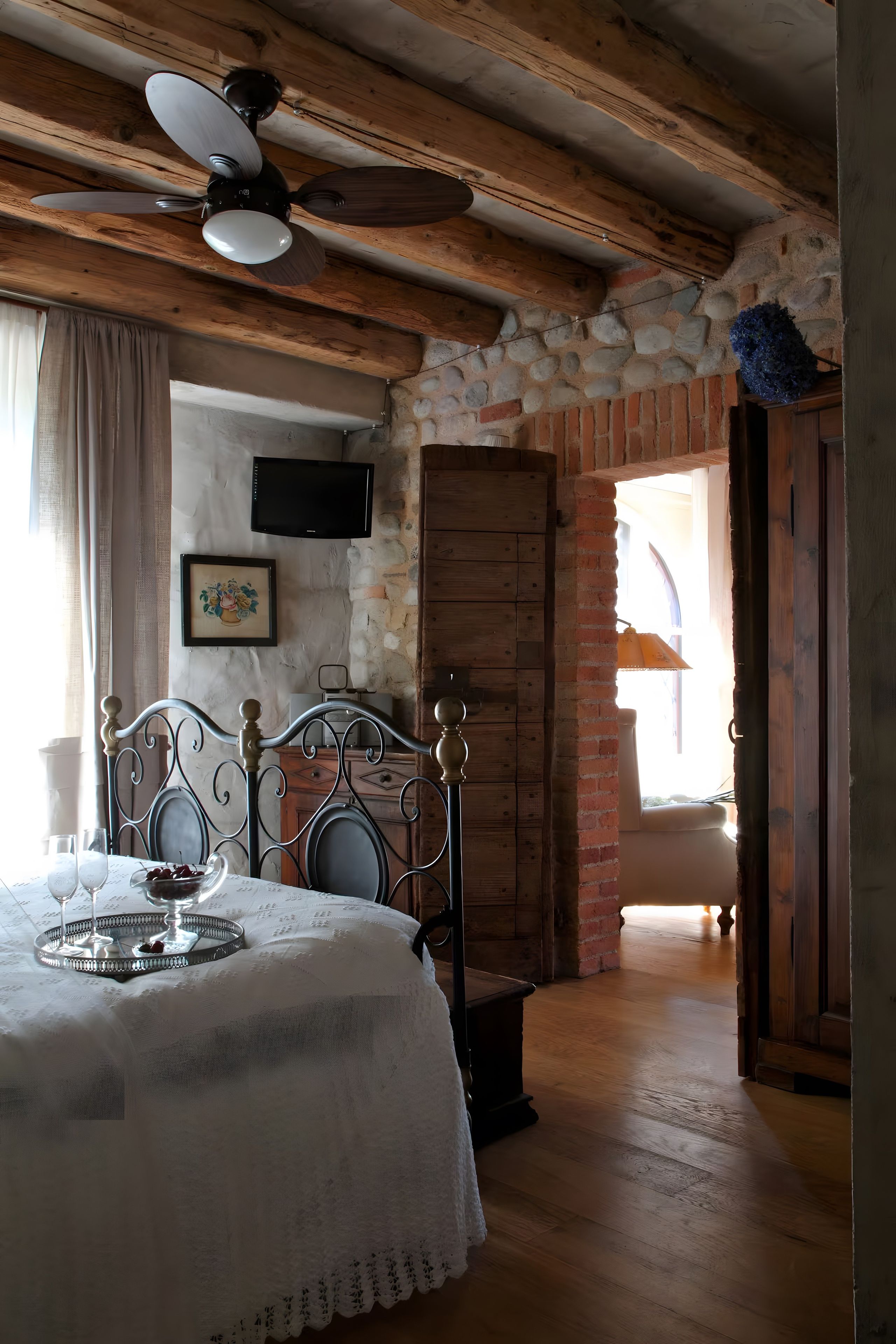 Ein Schlafzimmer mit einem Bett, einem Holzkopfteil, einer Steinwand, einem Deckenventilator und einem Fenster mit Vorhängen.