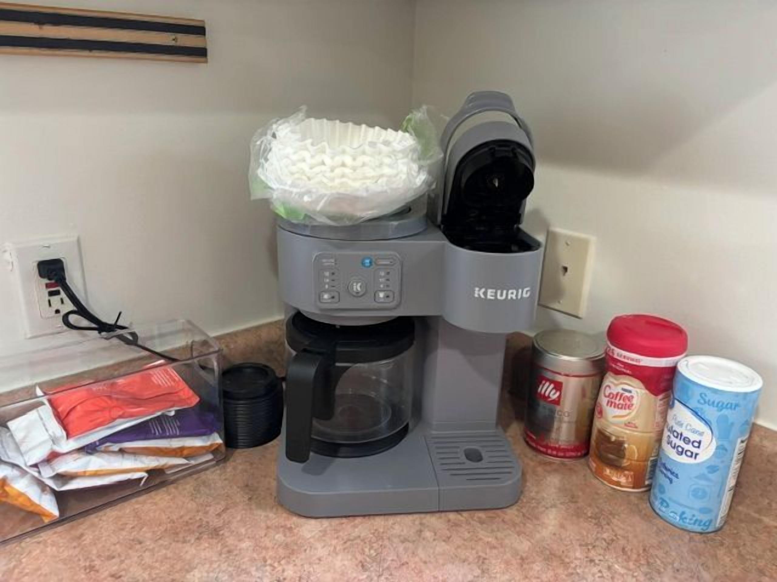 En kaffebryggare med en filterhållare, en burk kaffe, en burk socker och en behållare med grädde står på en bänkskiva.