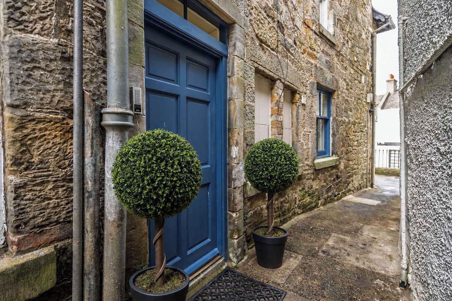 A stone building with a blue door and two potted topiary plants.