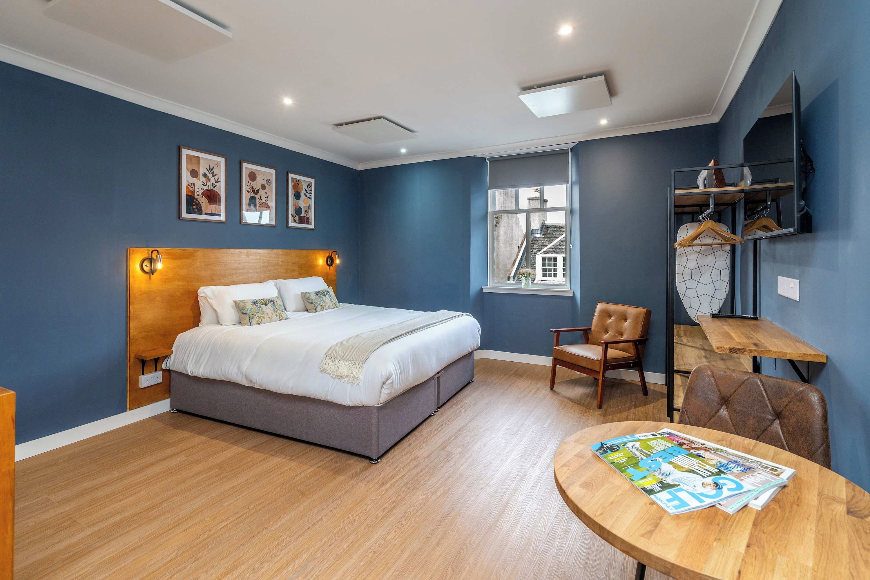 A modern bedroom with a large bed, a wooden headboard, a small table with magazines, a chair, and a window with a view of buildings.