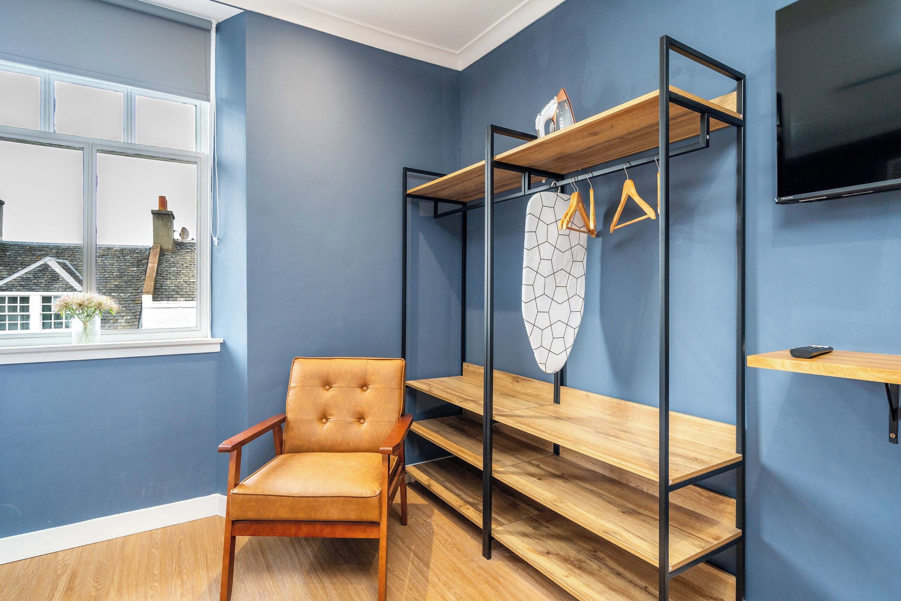 Una habitación con pared azul, una silla de madera, una cama con colchón blanco y un televisor montado en la pared.