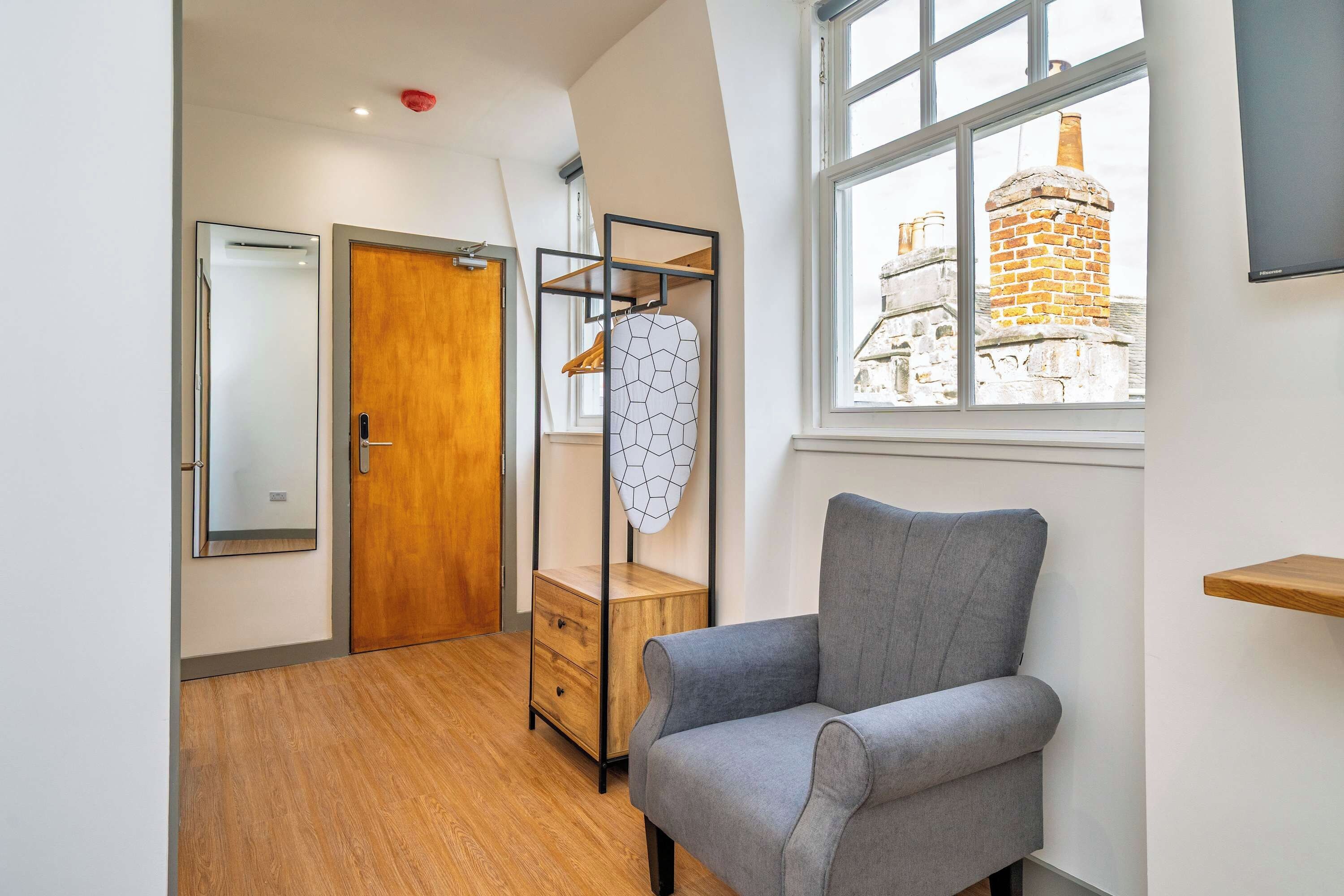 A modern room with a grey armchair, wooden wardrobe, and a window with a view of a brick building.