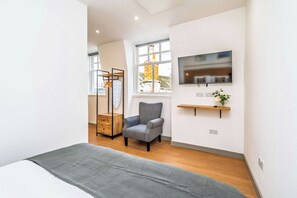 A modern bedroom with a grey sofa, a wooden dresser, a TV mounted on the wall, and a window with a view of a brick building. - Cozy Studio at The Brewery (Queensferry)