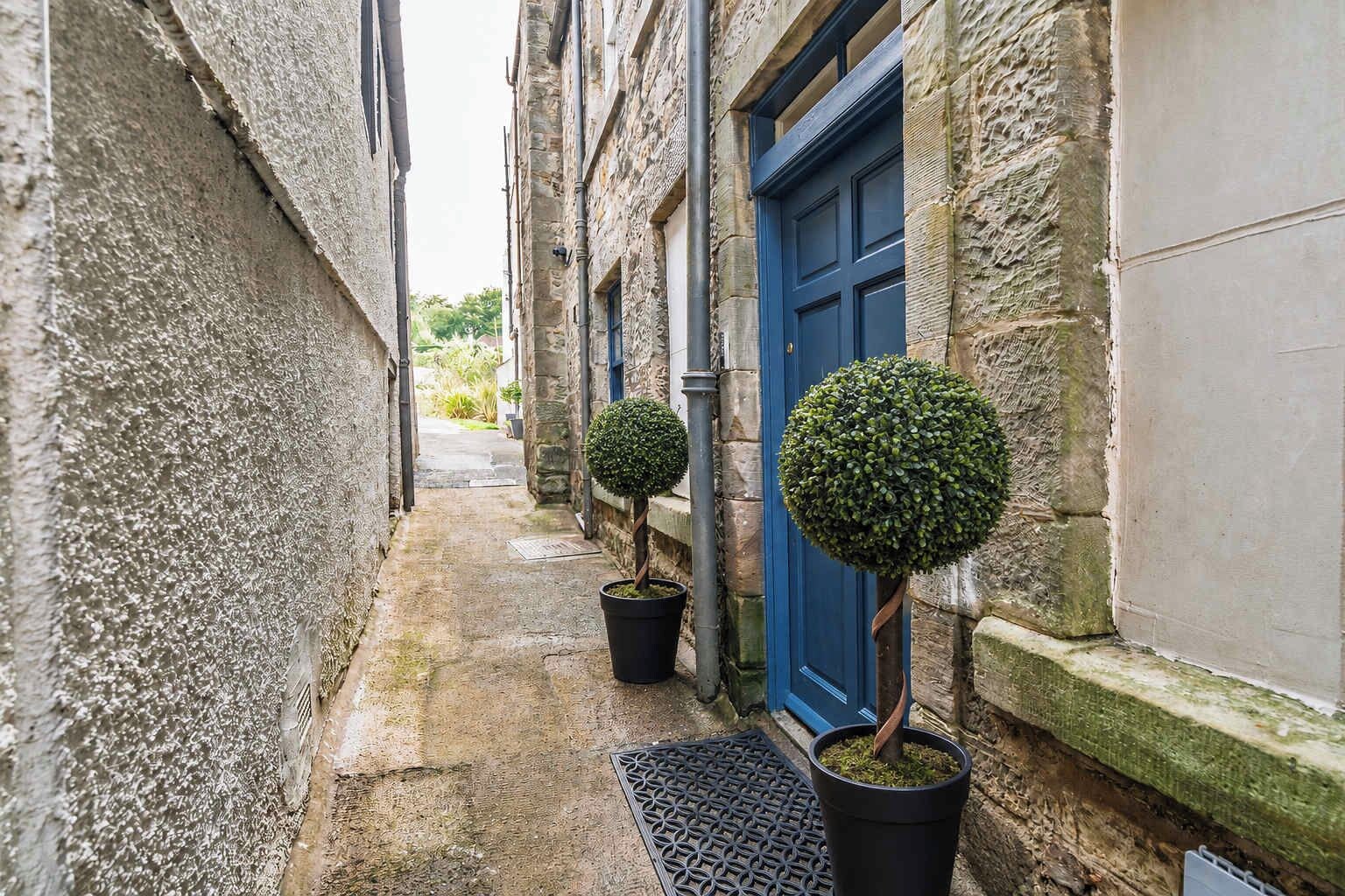A narrow alleyway with stone walls, a blue door, and two potted topiary trees.