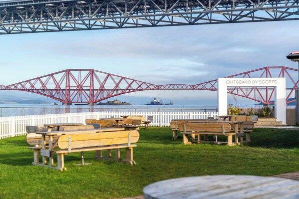 An outdoor seating area with wooden benches and tables, a white fence, and a sign that reads 'OUTBOARD BY SCOTTS'. - Cozy Boutique Studio at The Brewery (Queensferry)
