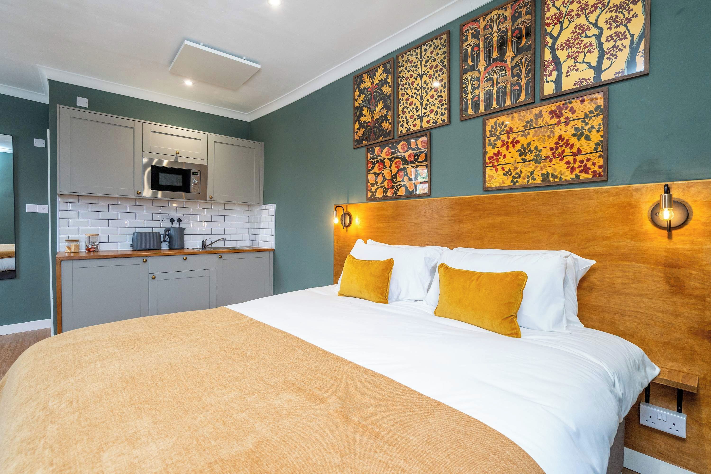 Une chambre à coucher comprenant un lit, deux oreillers, une tête de lit et un luminaire fixé au mur, ainsi qu’un coin cuisine équipé d’un four à micro-ondes.